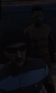 a man wearing sunglasses and a beanie stands next to another shirtless man