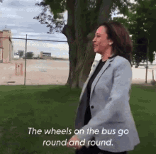 a woman in a grey jacket is walking in a park with the words " the wheels on the bus go round and round " above her