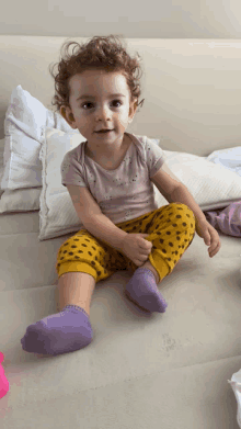 a little girl wearing purple socks and yellow pants sits on a bed