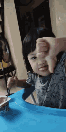 a little girl is sitting at a table eating yogurt with a spoon and giving a thumbs down sign .