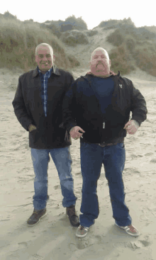 two men standing next to each other on a beach with one wearing a black jacket