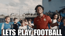 a man in a red shirt stands in front of a group of children with the words let 's play football above him