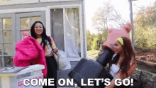 two women are standing next to each other in front of a house and one of them is holding a large suitcase .