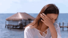 a woman is covering her face with her hand while standing on a balcony overlooking the ocean with a hut in the background .