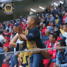 a boy is being carried by a man in a stadium with a pumas logo on the wall behind him