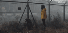 a woman in a yellow jacket stands in front of a barbed wire fence with netflix written in the corner