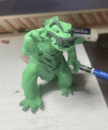 a green figurine of a lizard is sitting on a piece of graph paper next to a marker