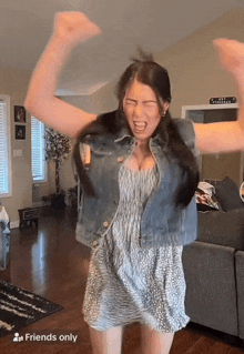 a woman in a dress and denim vest is dancing in a living room with her arms in the air