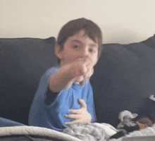 a young boy in a blue shirt is sitting on a black couch and pointing at the camera .