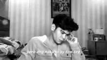 a black and white photo of a young man sitting on a bed with his hand on his head .