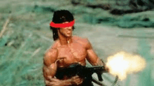 a man without a shirt is holding a gun with a red band around his head .