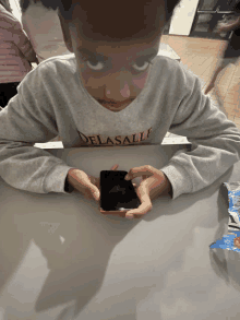 a young boy wearing a delasalle sweatshirt sits at a table looking at his phone