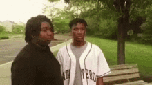 two men are standing next to each other on a sidewalk in a park . one of the men is wearing a baseball jersey .