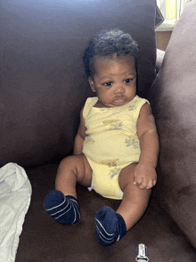 a baby is sitting on a brown couch wearing a yellow bodysuit with bananas on it