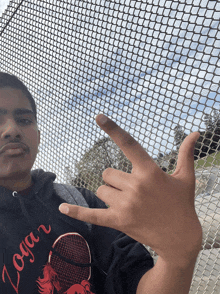 a young man wearing a jogar shirt makes a peace sign in front of a chain link fence