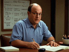 a man wearing glasses sits at a desk with a white board behind him