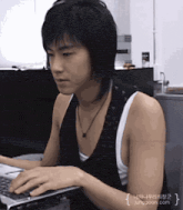 a young man is typing on a laptop computer while wearing a black vest and white tank top ..