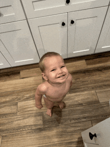 a baby without a shirt is standing on a wooden floor in front of white cabinets