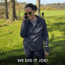 a woman wearing sunglasses is talking on a cell phone while standing in a grassy field .