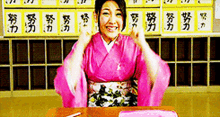 a woman in a pink kimono is sitting at a desk in front of a wall with chinese characters on it