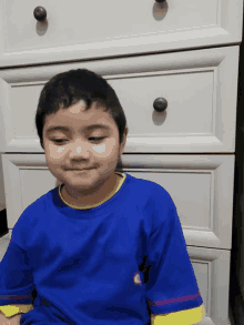 a young boy wearing a blue shirt with a yellow stripe on the sleeve is sitting in front of a white dresser