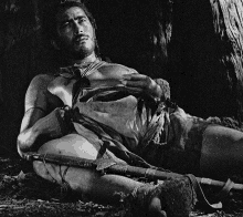 a black and white photo of a man laying on the ground with a sword in his hand .