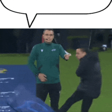 a man in a green jacket is pointing at another man on a soccer field