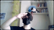 a man wearing a black mask salutes in front of a shelf full of books