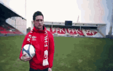 a man in a red jacket holds a soccer ball in front of a stadium with ffk written on it