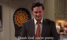 a man in a suit and tie is standing in front of a dart board and says `` thank god you 're pretty ''