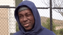 a man wearing a blue hoodie winks at the camera while standing in front of a chain link fence .