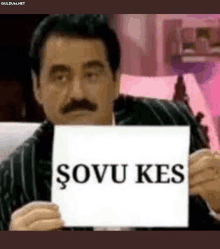 a man with a mustache is holding a piece of paper that says sovun kes .