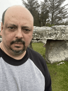 a bald man with a beard and mustache stands in front of a large rock