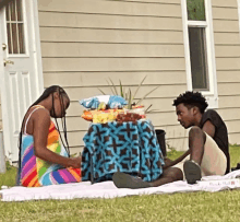 a woman in a colorful dress sits next to a man on a blanket in the grass