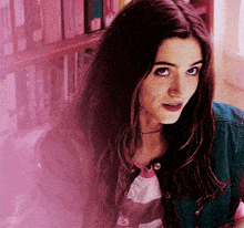 a woman in a denim jacket stands in front of a bookshelf and looks at the camera