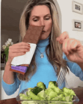 a woman in a blue sweater is eating broccoli and a bar of milka chocolate