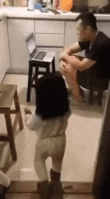 a little girl is standing in front of a laptop while a man sits on a stool in the kitchen .