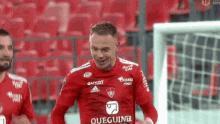 a soccer player wearing a red jersey with the word queguiner on it is standing in front of a soccer goal .