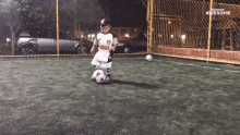 a young boy is kicking a soccer ball on a field with a fence in the background that says awesome