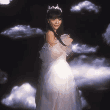 a woman in a white dress and tiara is standing in front of clouds