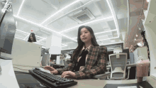 a woman in a plaid jacket sits at a desk in front of a computer with the twice logo on the ceiling