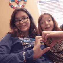 two girls are making a heart shape with their hands in front of a window with a sign that says " welcome " on it