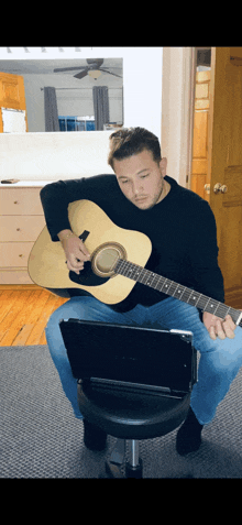 a man sits on a stool playing an acoustic guitar in front of a laptop