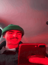 a man wearing a green beanie and a mustache is looking at a cell phone .