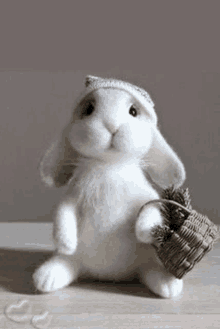 a white stuffed rabbit wearing a hat is holding a basket with a pine cone in it .