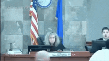 a woman is sitting at a desk in front of a computer in a courtroom with a flag in the background .