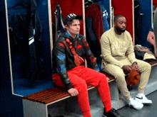 two men are sitting on benches in a locker room . one of the men is wearing a hat .