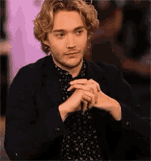 a young man with blonde hair is sitting at a table with his hands folded and looking at the camera .