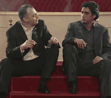 two men in suits are sitting on red steps smoking cigarettes