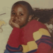 a young boy with his hand on his chin wearing a red yellow and blue sweater